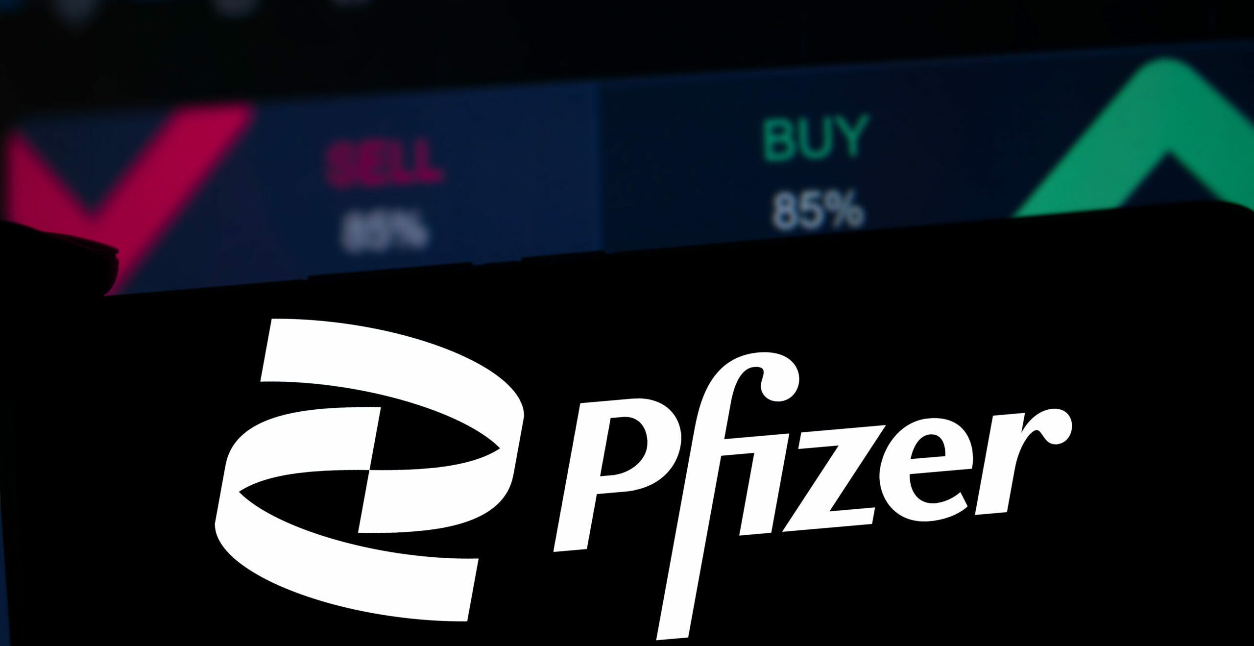 Symbolbild: Logo der Firma Pfizer auf einem Aktienhintergund. Worte wie Buy und Sell sind zu sehen.