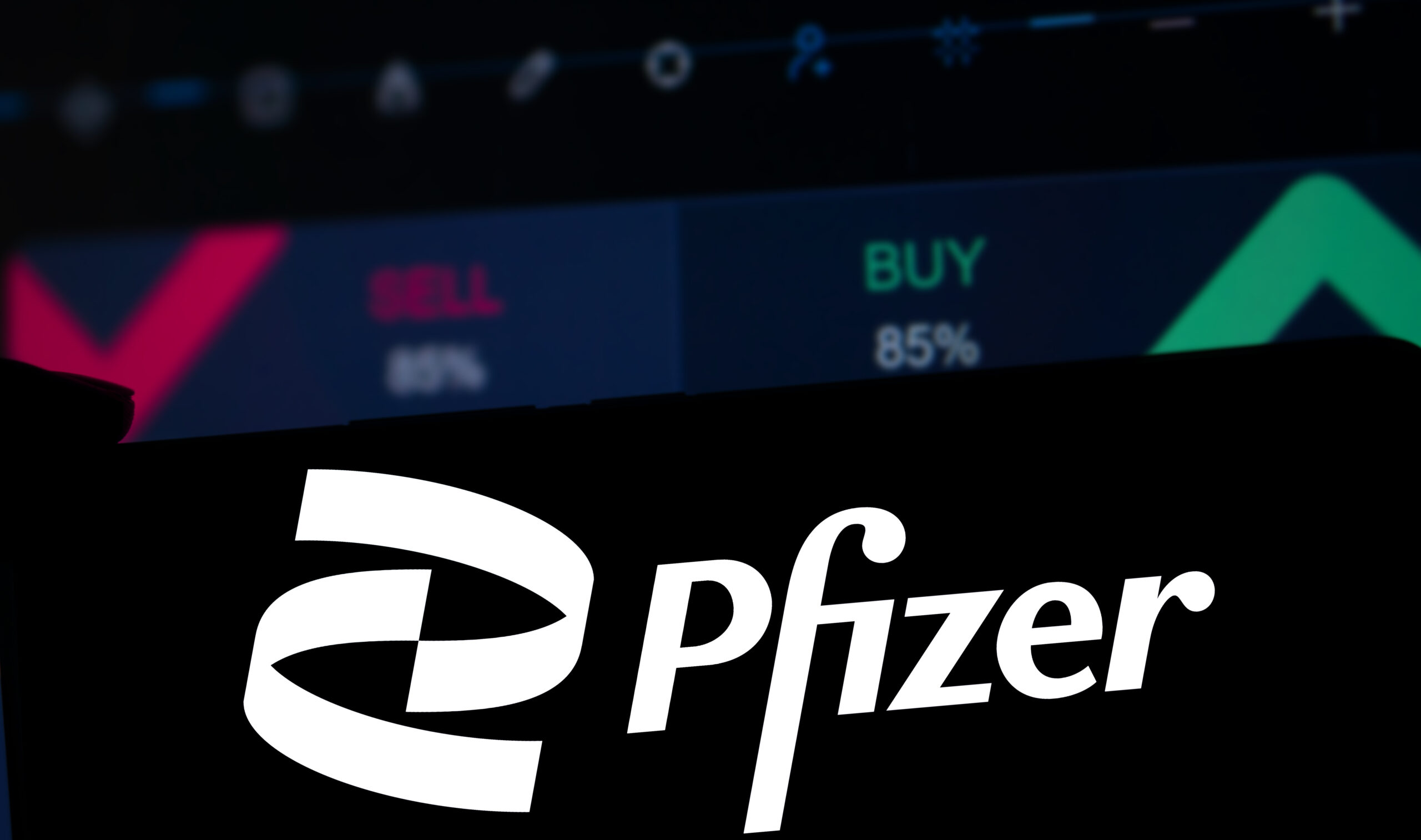 Symbolbild: Logo der Firma Pfizer auf einem Aktienhintergund. Worte wie Buy und Sell sind zu sehen.