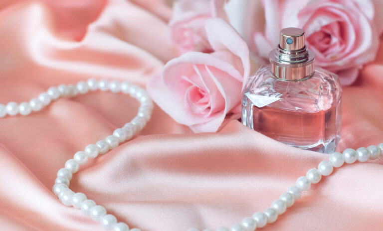 Symbolbild: Auf einem rosa Stoff liegt eine Perlenkette, ein Parfümfläschchen und eine Rose