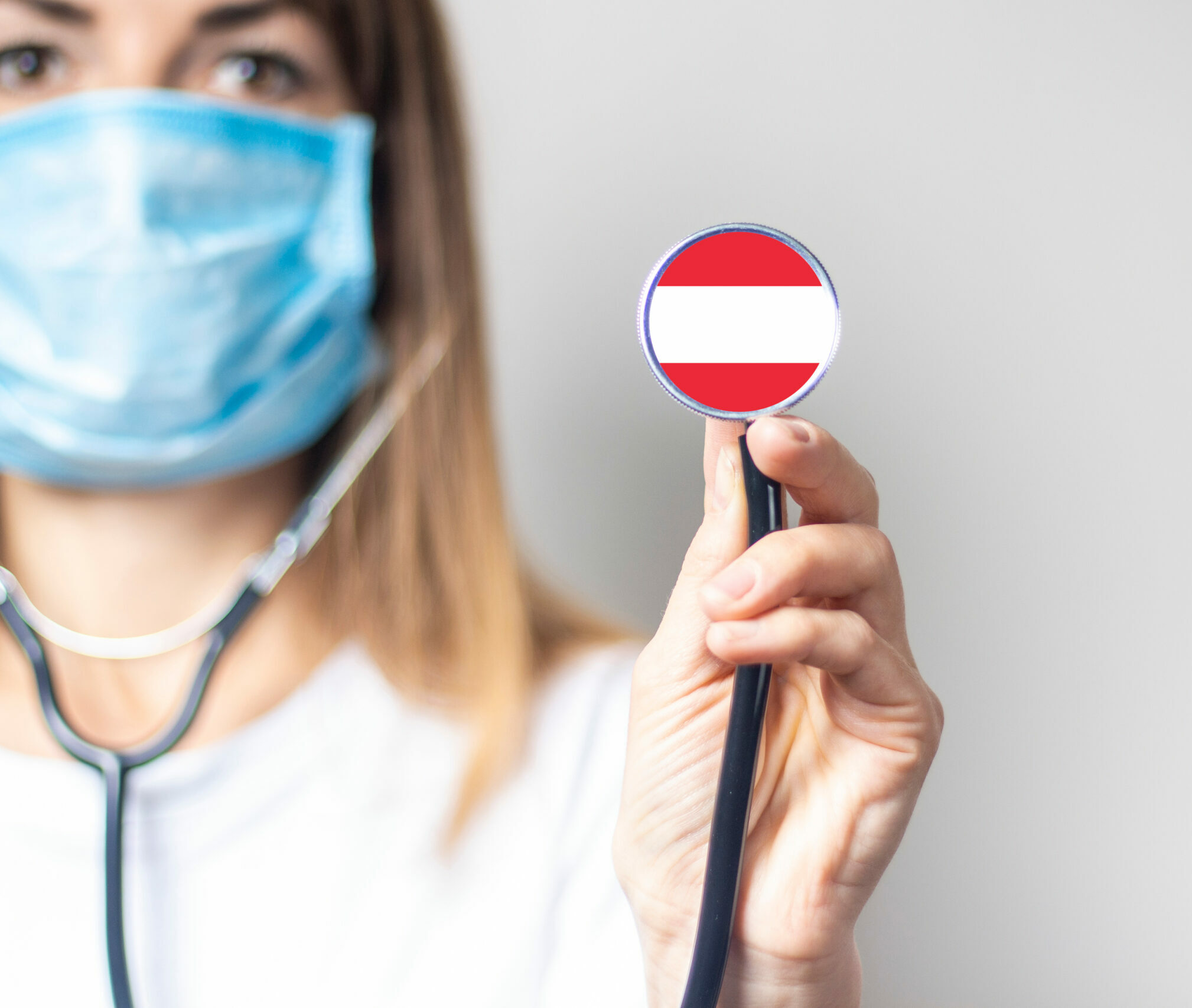 Symbolbild: Eine Frau hat eine Maske auf und hält ein Stethoskop hoch. Dieses trägt die Rot-Weiß-Rote Flagge von Österreich.