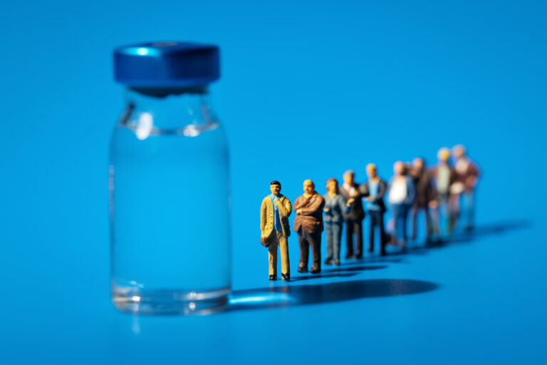 Symbolbild: Ein Impfstofffläschchen mit einer Reihe von Plastikfiguren. Sie stellen sich dahinter an und warten symbolisch auf die Impfung.