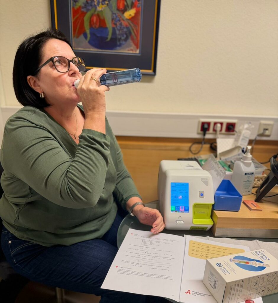 Mitarbeiterin Priska sitz im Testraum und zeigt die Atemmessung vor. Sie hält das Diagnosegerät in der Hand und führt es an ihre Lippen.