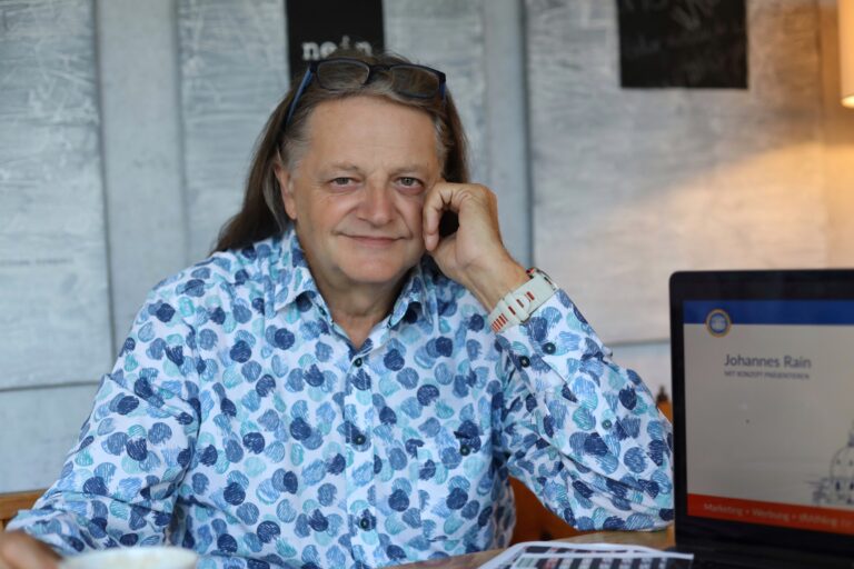 Hannes Rain sitzt in einem Hemd mit blauen Kreisen an einem Tisch. er hält die Hand an die Wange und lächelt. Neben ihm steht ein Laptop.