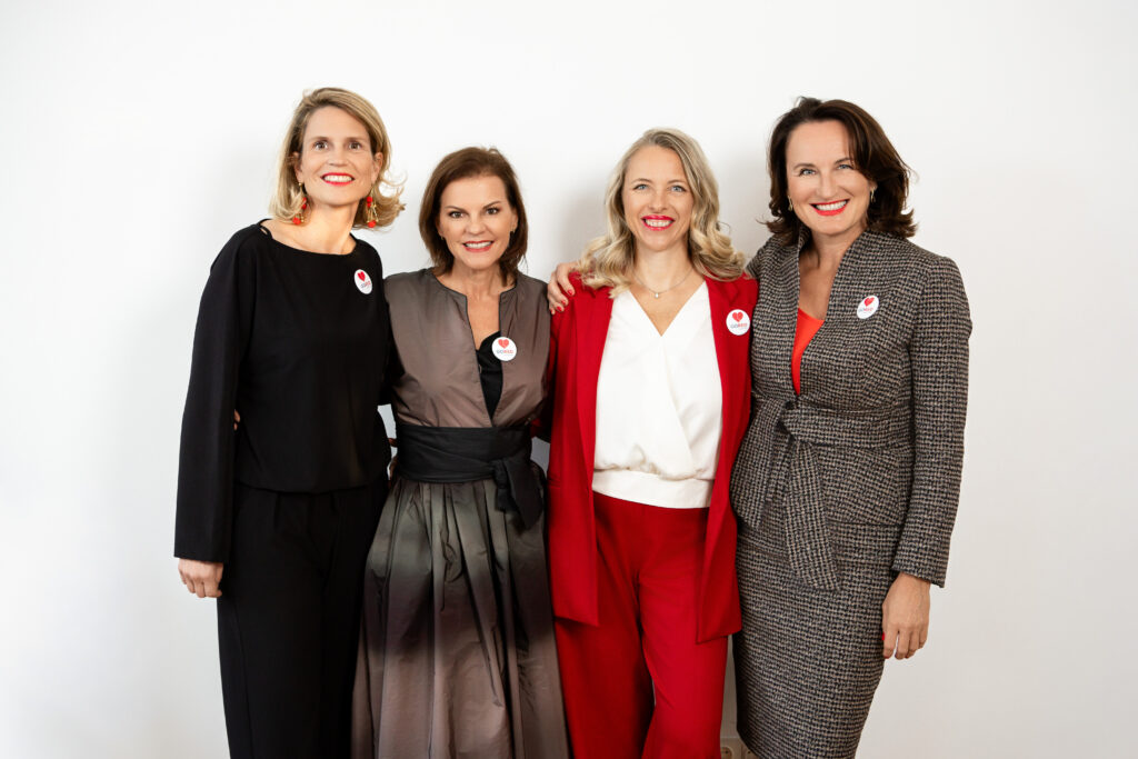 Die vier Damen sind die Initiatorinnen von GoRed Austria. Sie stehen Schulter an Schulter und lachen. Jede von ihnen trägt ein rotes Accessoire.