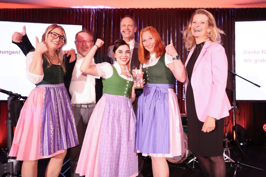 Die Gewinner:innen des Awards lächeln in die Kamera. Sie tragen Dirndl und werfen die Hände in die Höhe.