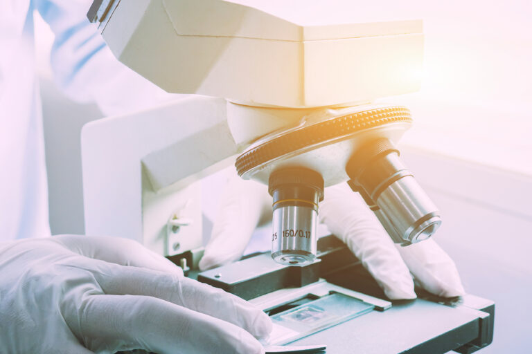 Symbolbild: Ein Mikroskop mit Händen eines Wissenschaftlers mit Handschuhen.