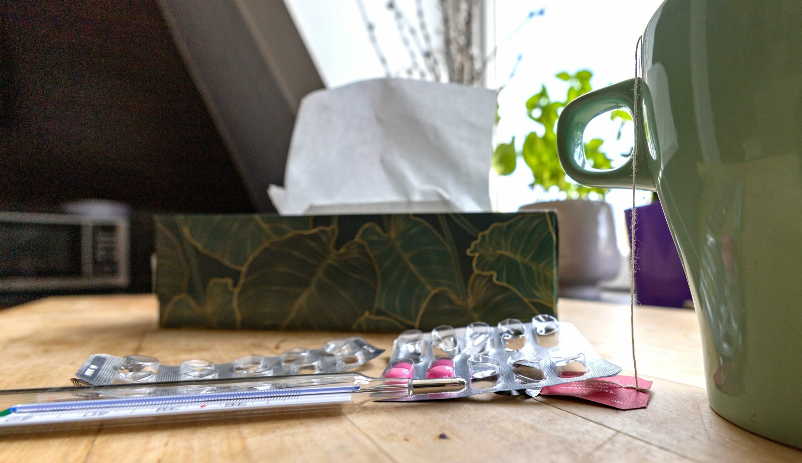 Symbolbild: Eine Tasse, ein Blinder mit pinken Tabletten, ein Fieberthermometer stehen an einem Tisch.