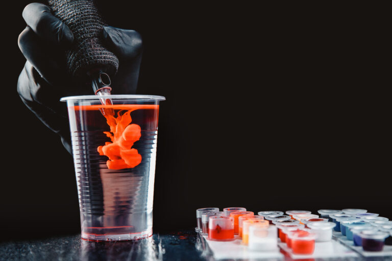 Symbolbild: Nahaufnahme einer Tätowiermaschine, bei der die Nadeln mit klarem Wasser von Tätowierpigmenten gereinigt werden.