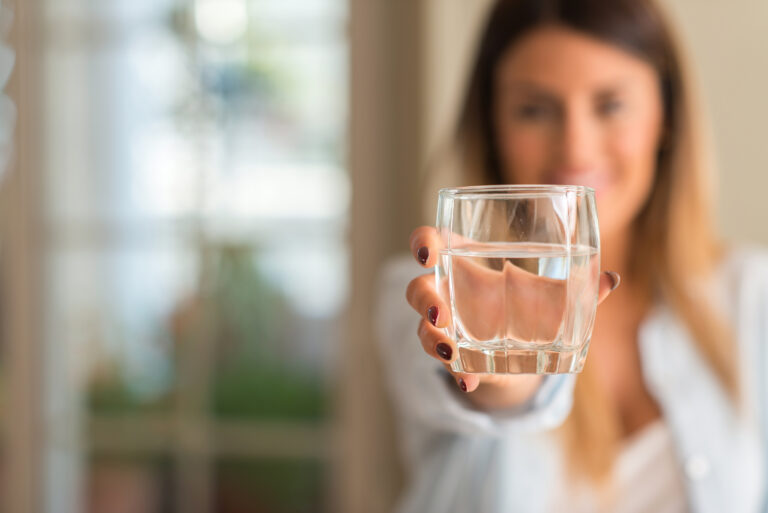 Symbolbild: Eine Junge Frau hält ein Wasserglas vor sich hin. Sie steht dahinter und ist verschwommen.