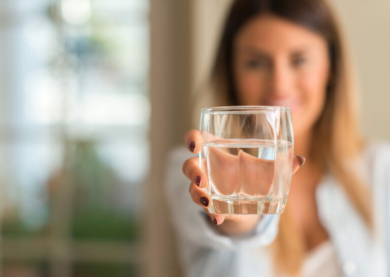 Symbolbild: Eine Junge Frau hält ein Wasserglas vor sich hin. Sie steht dahinter und ist verschwommen.