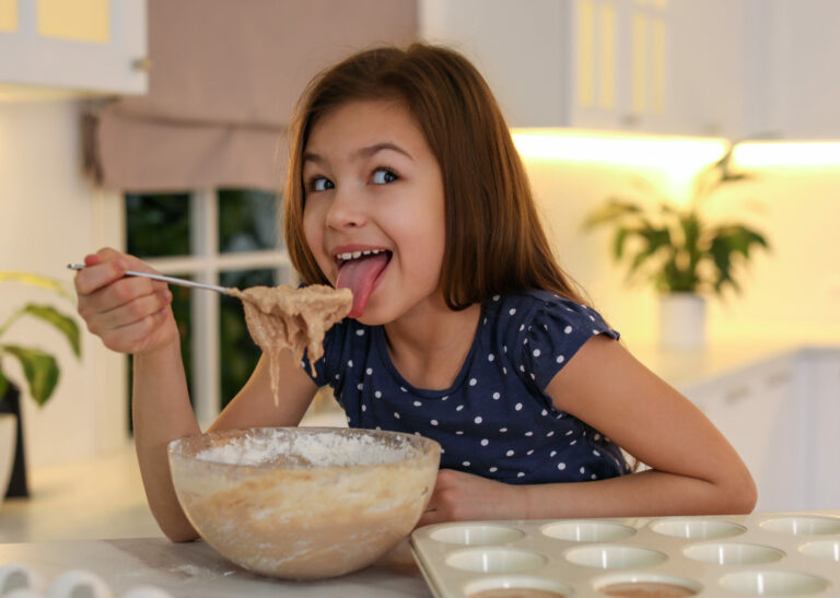 Symbolbild: Junges Mädchen schleckt rohen Keksteig von einem Kochlöffel. Neben ihr stehen Muffinförmchen.