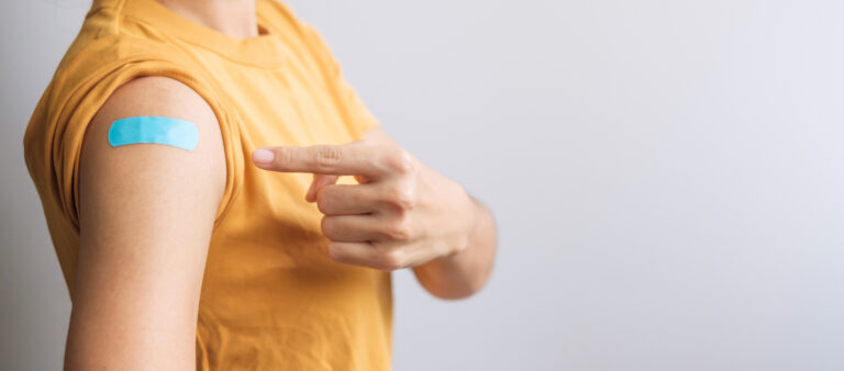 Symbolbild: Eine Frau im gelben Shirt zieht auf ihren Oberarm. Dort klebt ein Pflaster vom Impfen.
