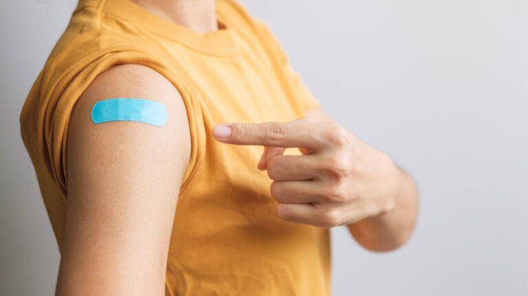 Symbolbild: Eine Frau im gelben Shirt zieht auf ihren Oberarm. Dort klebt ein Pflaster vom Impfen.