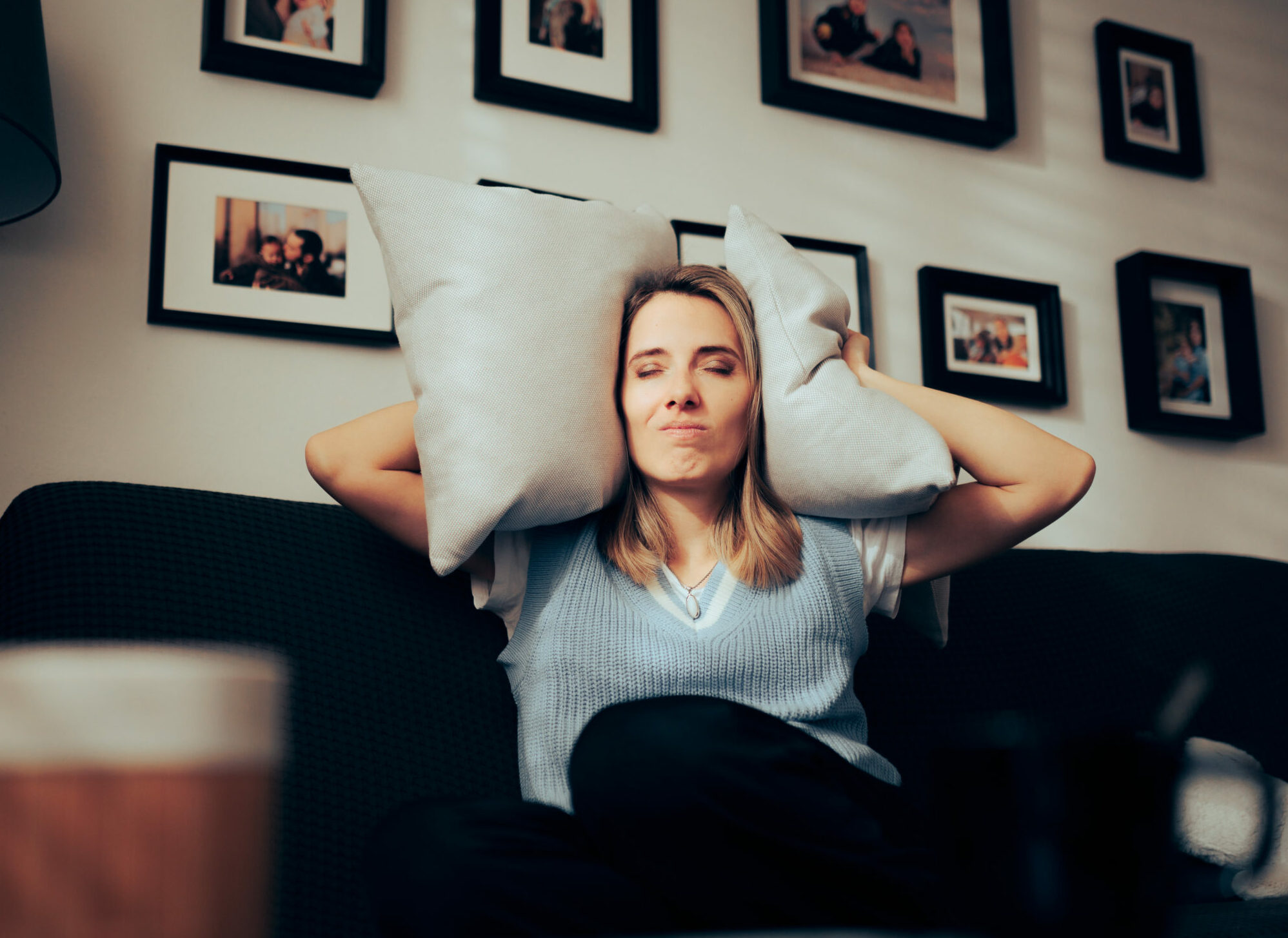 Symbolbild: Eine Frau sitz auf ihrem Sofa und hält sich Polster an die Ohren. Sie will den Lärm ausblenden.