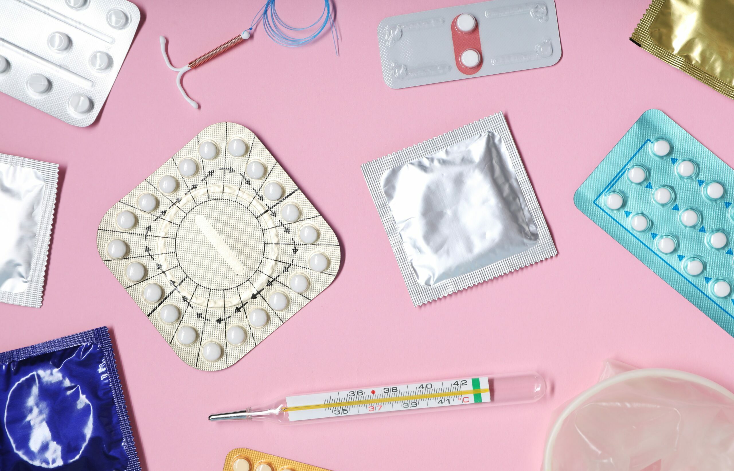 Symbolbdil: Auf einem rosafarbenen Hintergrund sind verschiedene Verhütungsmittel zu sehen. Eine Kupfer-Spirale, die Pille, Kondome und ein Thermometer.