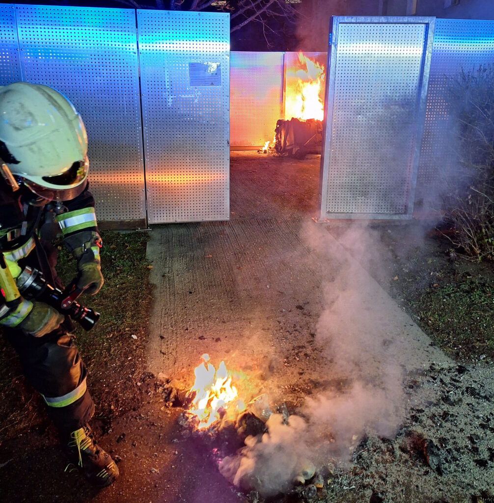 Die Freiwillige Feuerwehr löscht einen Brand. Dieser wurde durch eine Rakete ausgelöst. Der Feuerwehrmann hält einen Schlauch und zielt auf einen Mistkübel.