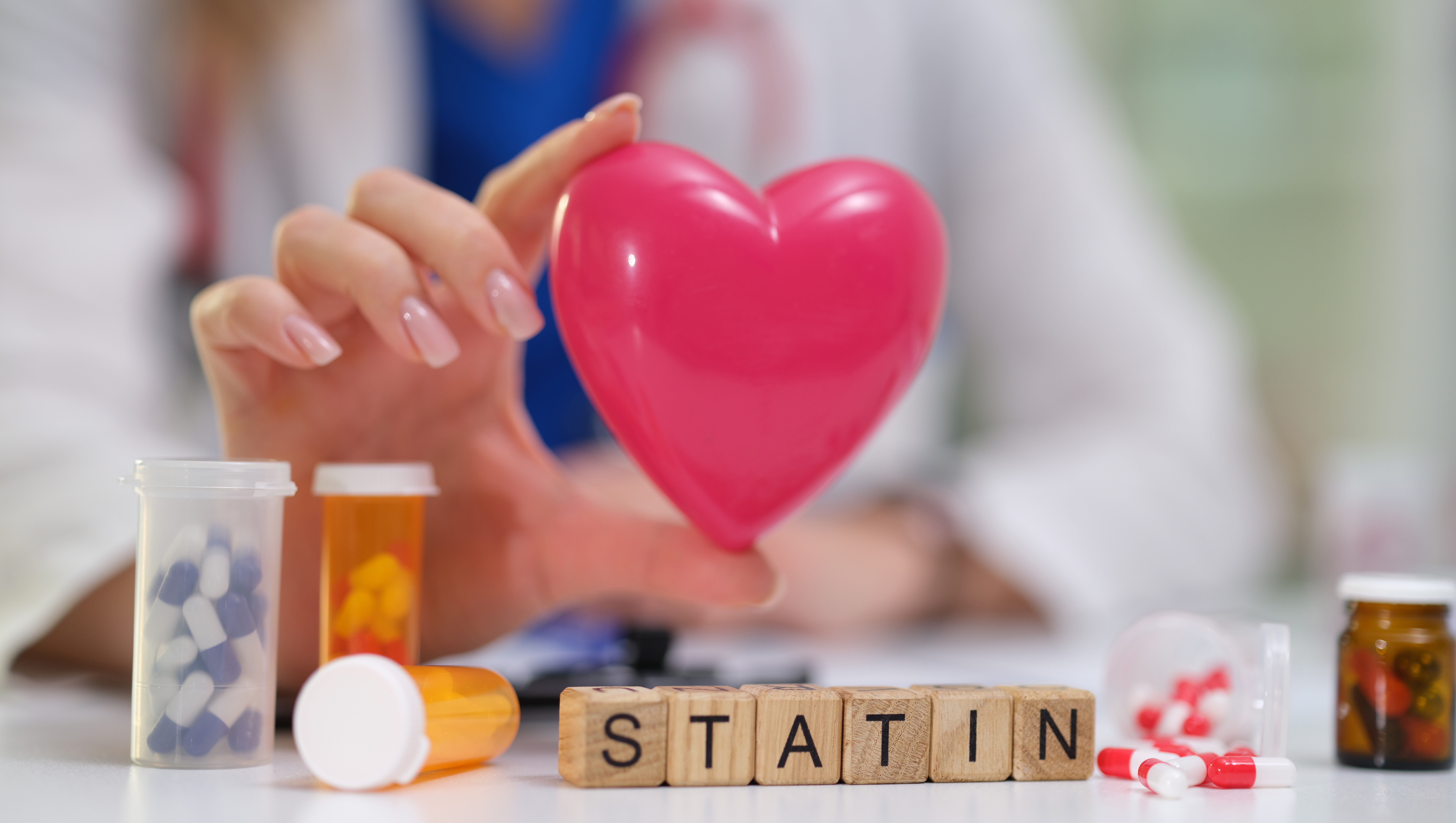 Symbolbild: Eine Hand hält ein Plastikherz. Es liegen Würfel mit der Aufschrift Statin herum und überall Kapseln.