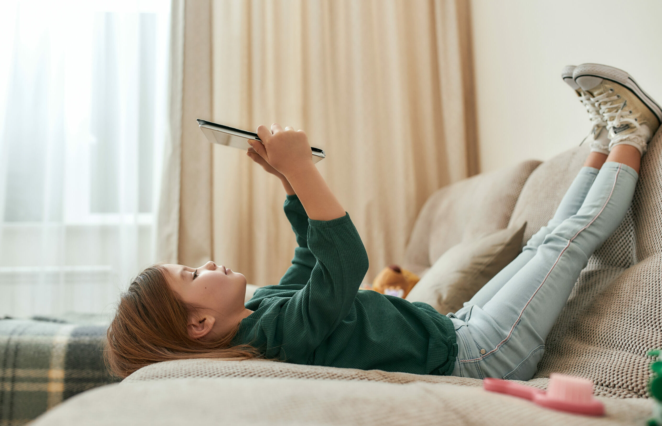 Symbolbild: Kleines Mädchen liegt am Sofa und hält ein Tablet vor sich in die Höhe.