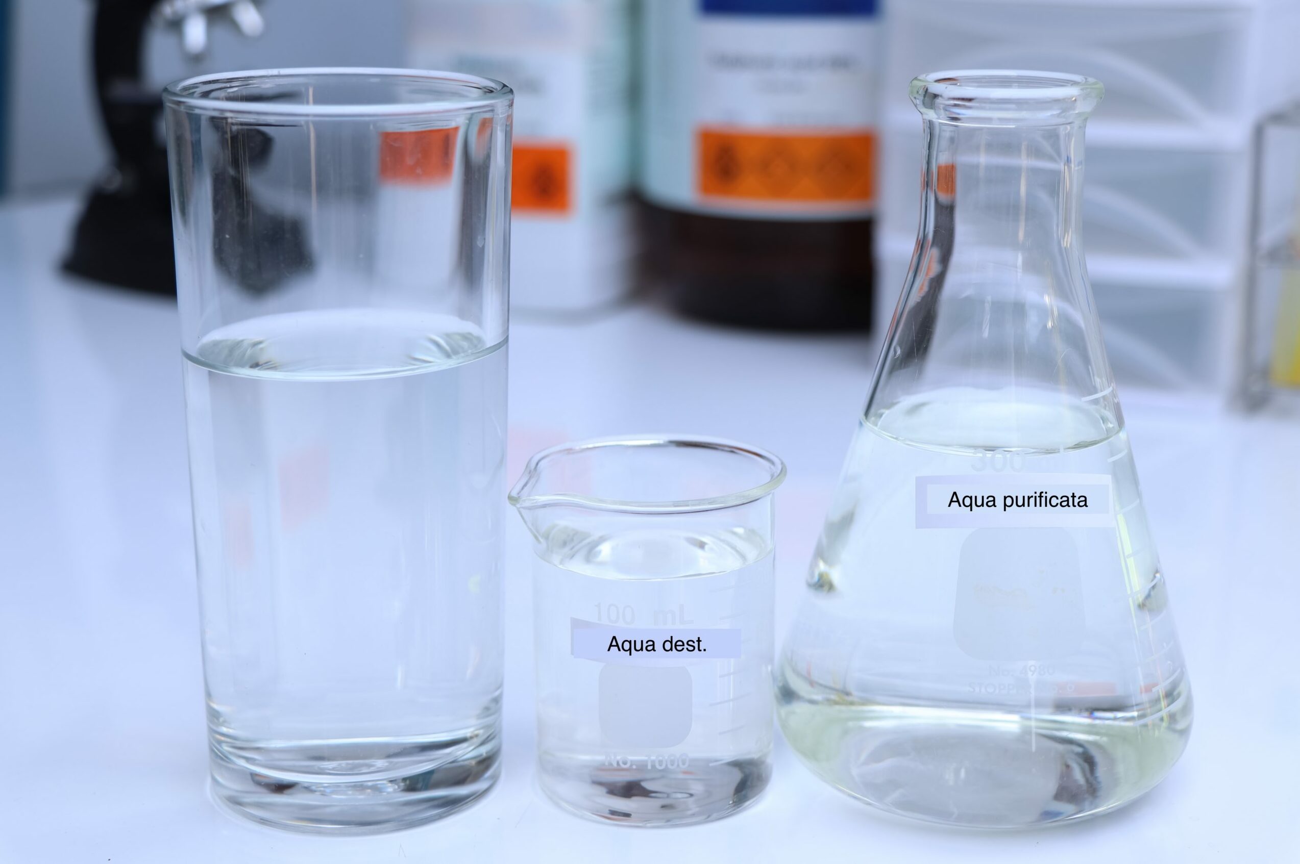 Symbolbild: Drei Glasgeräte im Labor mit Wasser gefüllt. Die Etiketten kennzeichnen es als Aqua Dest und Aqua Purificata.