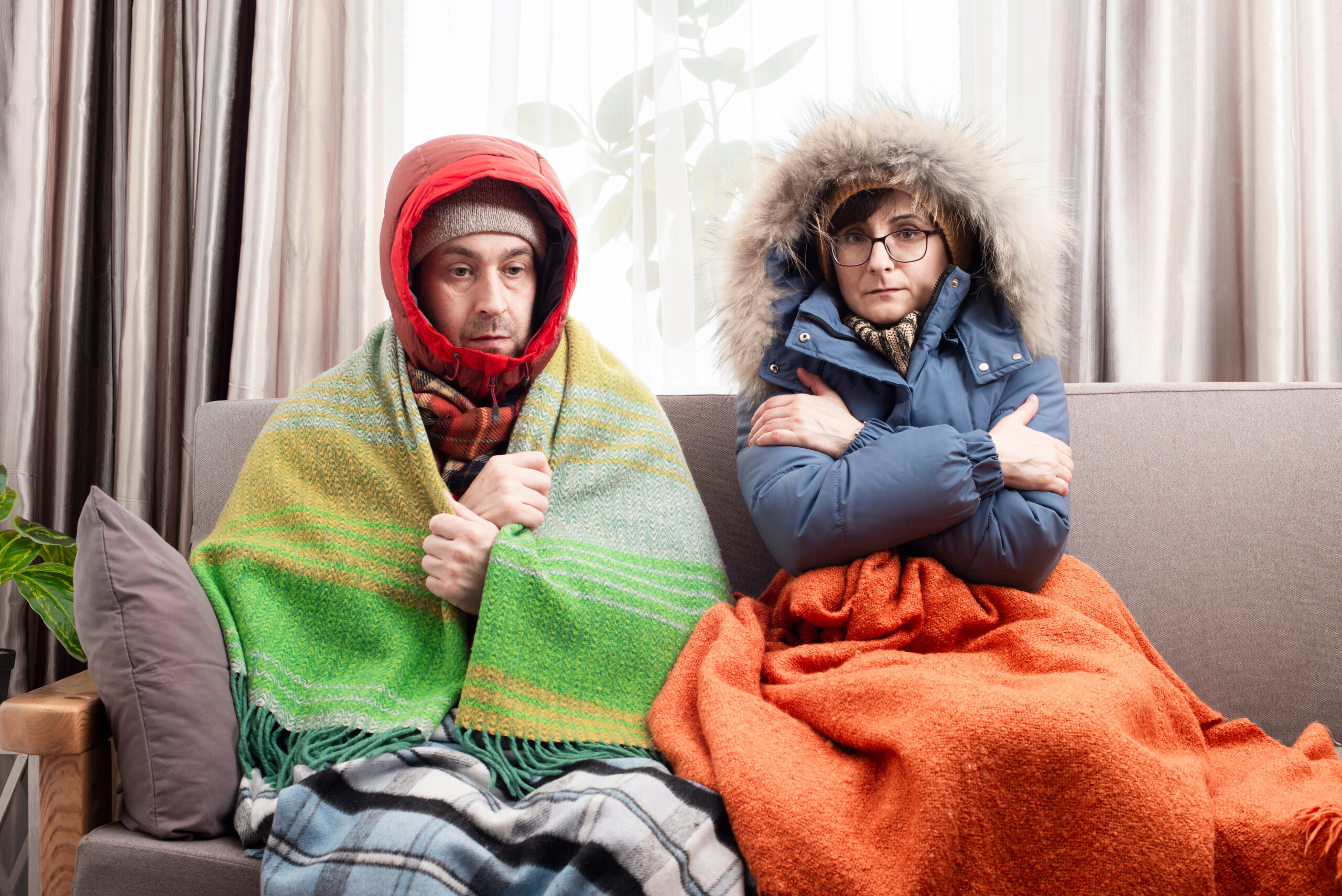 Symbolbild: Trauriges Ehepaar mittleren Alters in warmer Kleidung, das frierend auf dem Sofa in seinem Wohnzimmer sitzt.