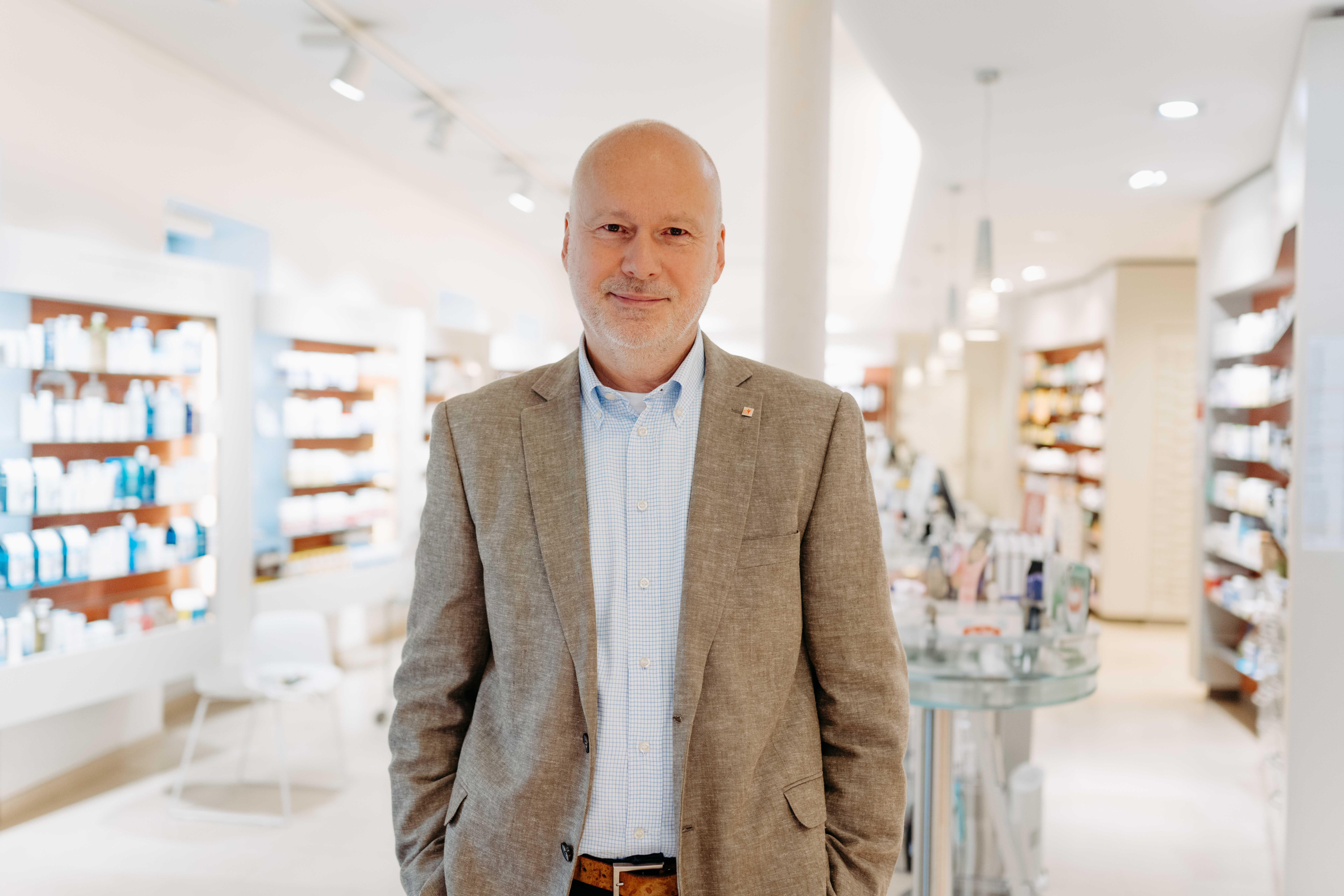 Mag. Geisler steht in einer Apotheke. Er trägt einen beigen Anzug.