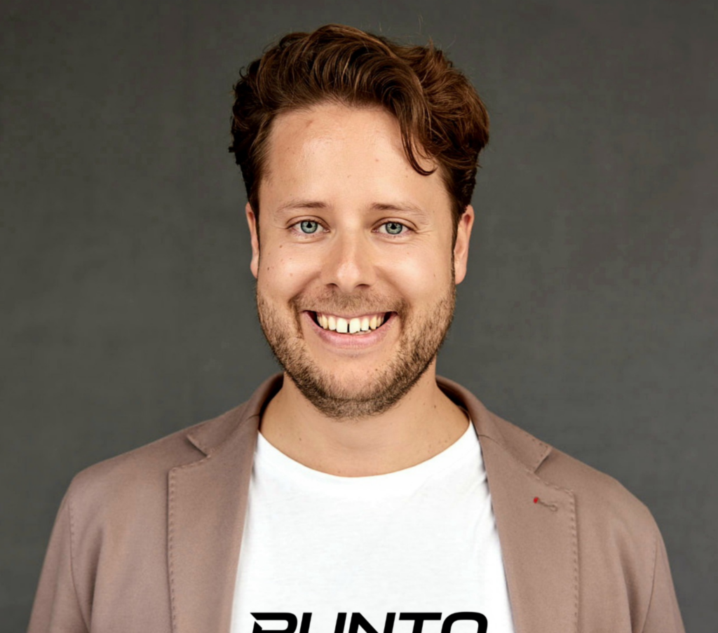 Maximilian Milenkovics trägt ein weißes Shirt und eine braunes Jackett. Er lächelt.