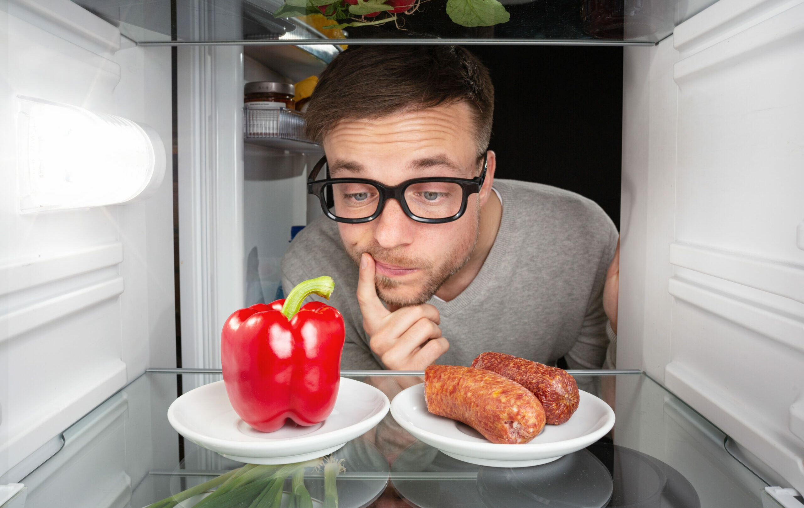Symbolbild: Mann schaut in Kühlschrank und will zwischen Paprika und Wurst entscheiden.