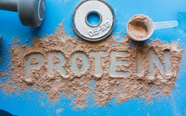 Sybolbild: In Proteinpulver wurden die Worte "Protein" geschrieben. Daneben liegen eine Hantel und eine Trainingsscheibe.