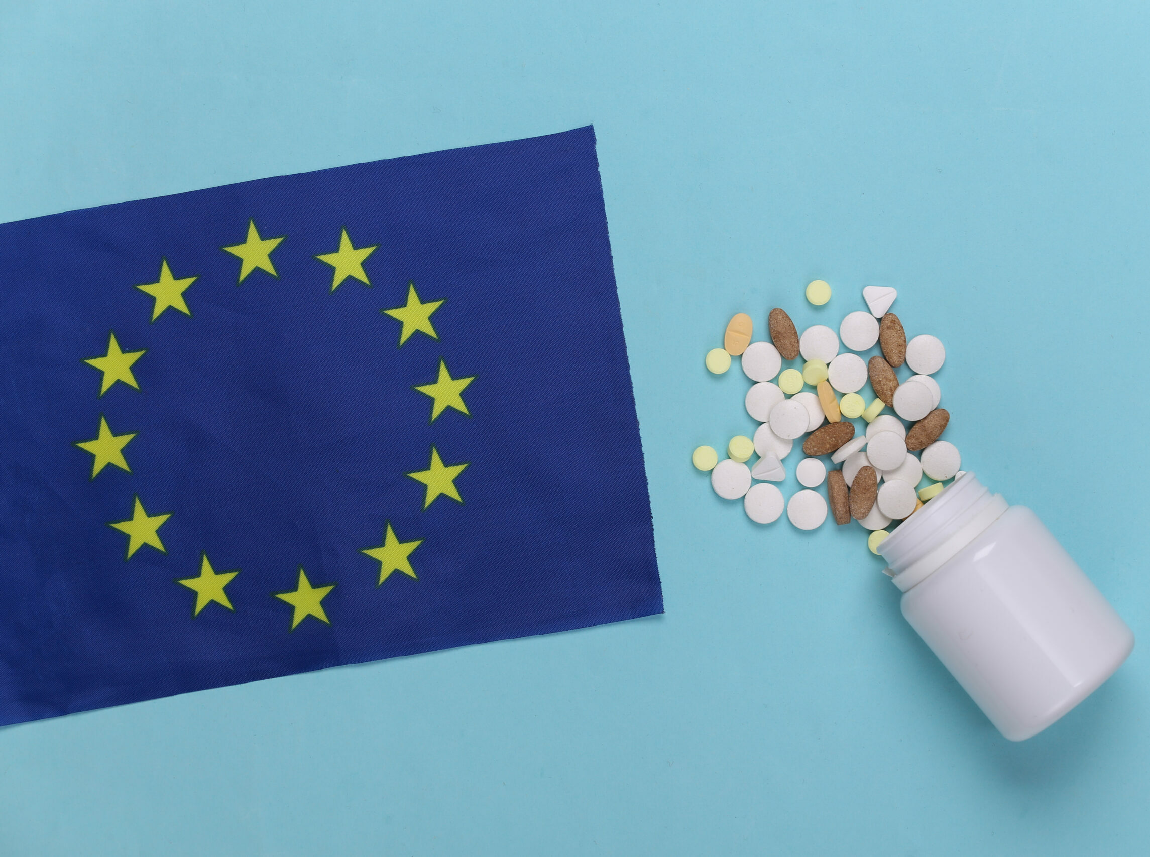 Symbolbild: Auf einem hellblauen Hintergrund liegt eine ausgeschüttete Pillen-Dose. Viele Tabletten und Pillen liegen neben einer blauen EU-Fahne.