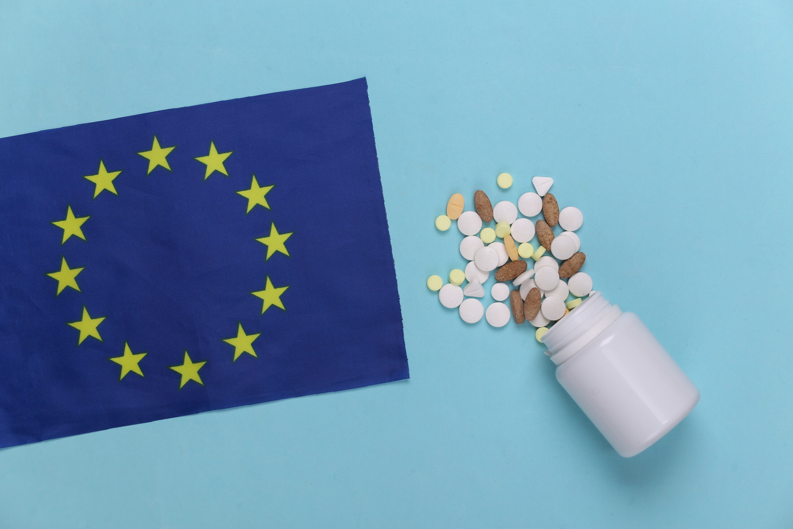 Symbolbild: Auf einem hellblauen Hintergrund liegt eine ausgeschüttete Pillen-Dose. Viele Tabletten und Pillen liegen neben einer blauen EU-Fahne.