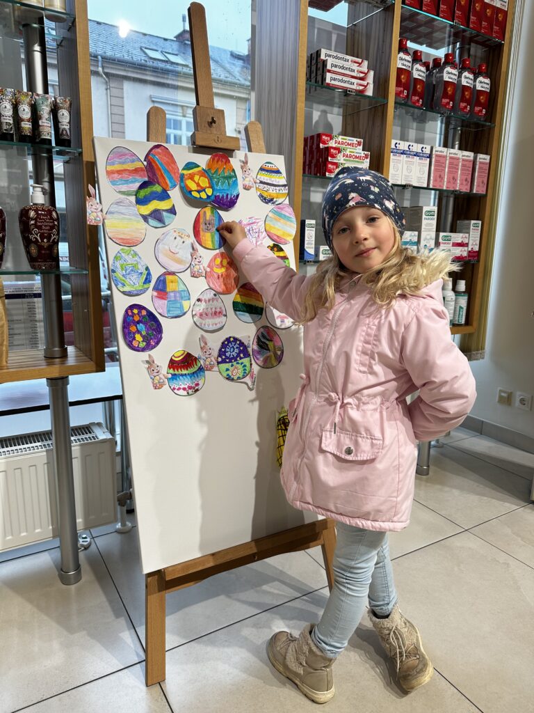 Stolz und mit einem lächeln präsentiert ein Kind sein Kunstwerk an der farbenfrohen Oster Collage in der Apotheke zur Heiligen Dreifaltigkeit in Berndorf.