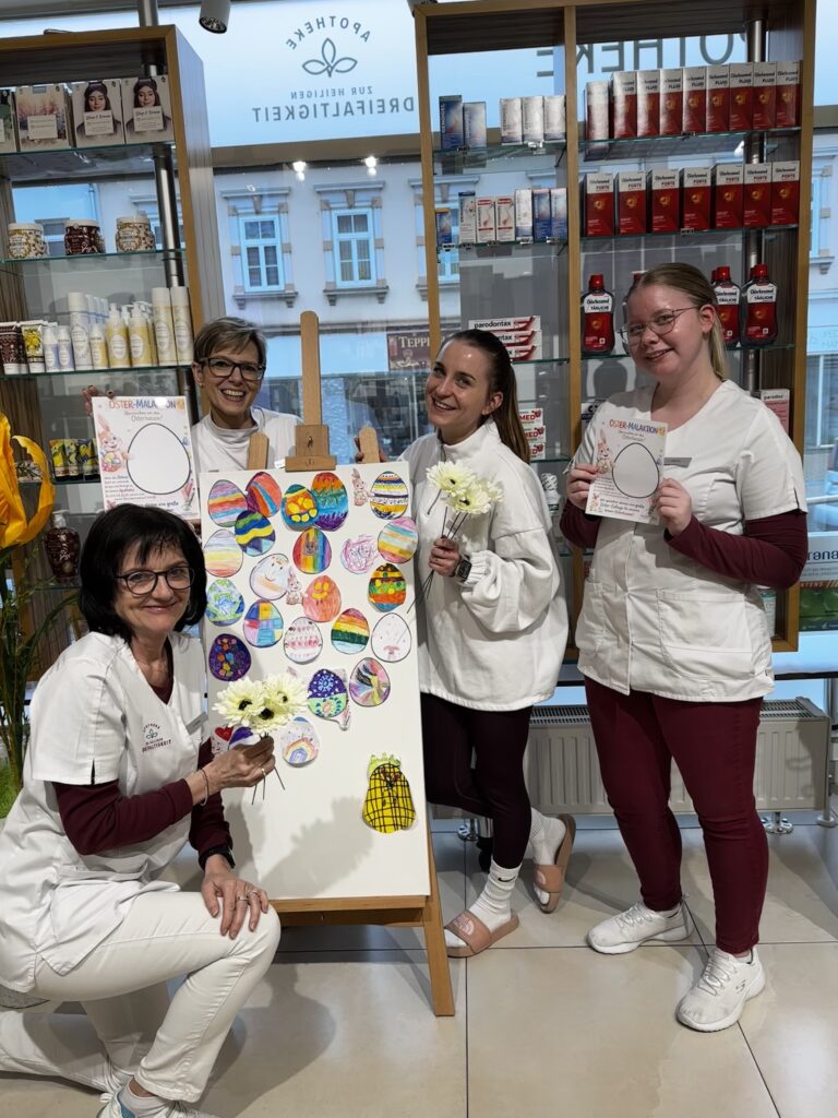 Apothekenteam der Apotheke zur Heiligen Dreifaltigkeit in Berndorf präsentiert eine bunte Collage aus von Kindern gestalteten Ostereiern im Schaufensterbereich der Apotheke.