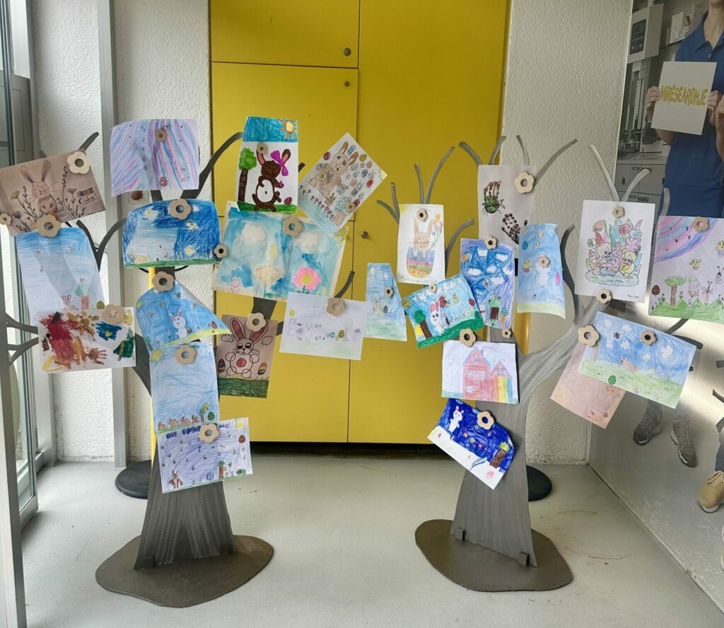 Eingangsbereich einer Apotheke mit zwei dekorativen Bäumen, an denen zahlreiche bunte Kinderzeichnungen mit Ostermotiven befestigt sind, vor einer gelben Wand.