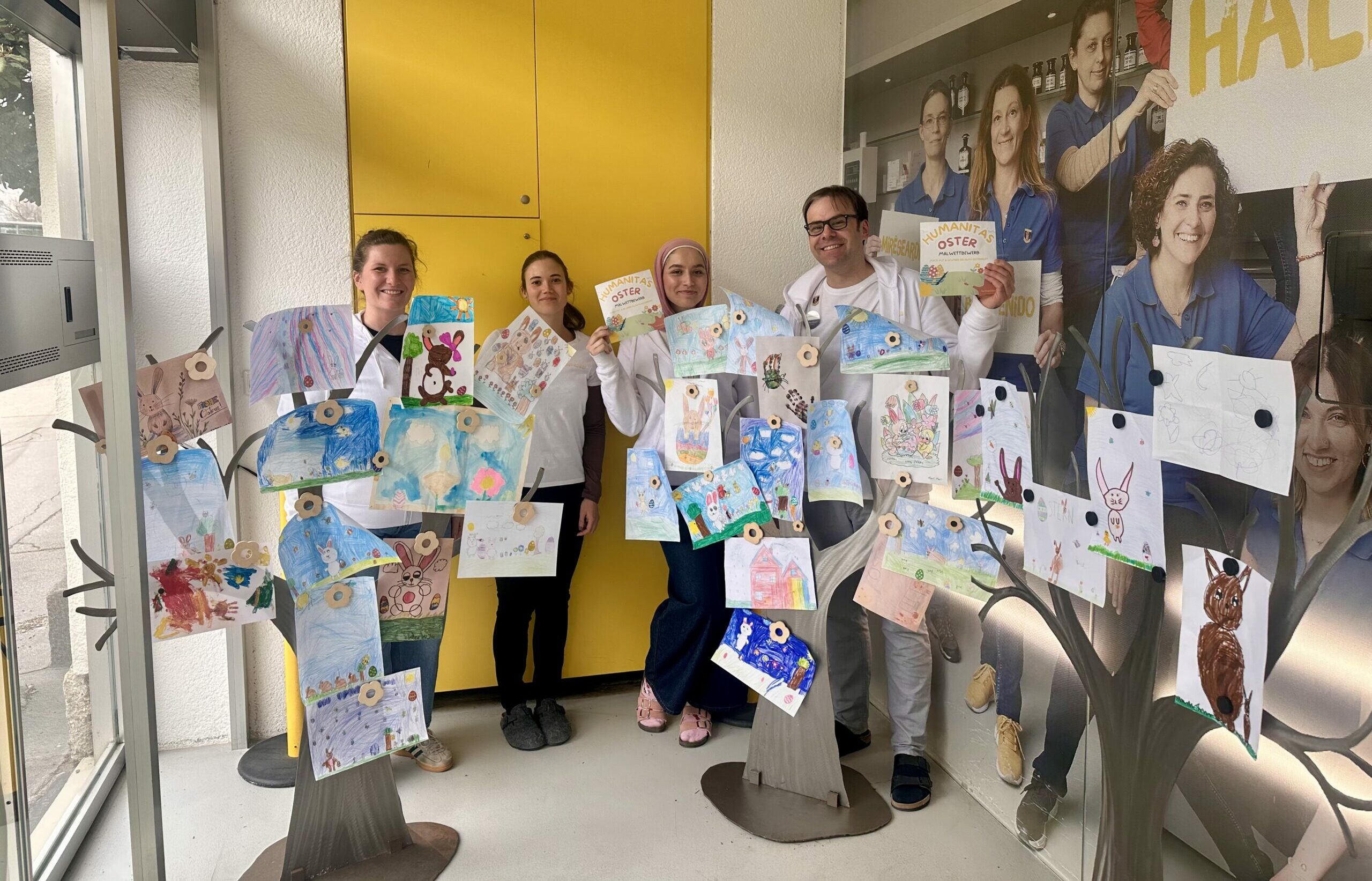 Das Apotheken Team präsentiert eine Oster-Kinderaktion mit bunten Zeichnungen von Kindern, die auf dekorativen Bäumen befestigt sind.