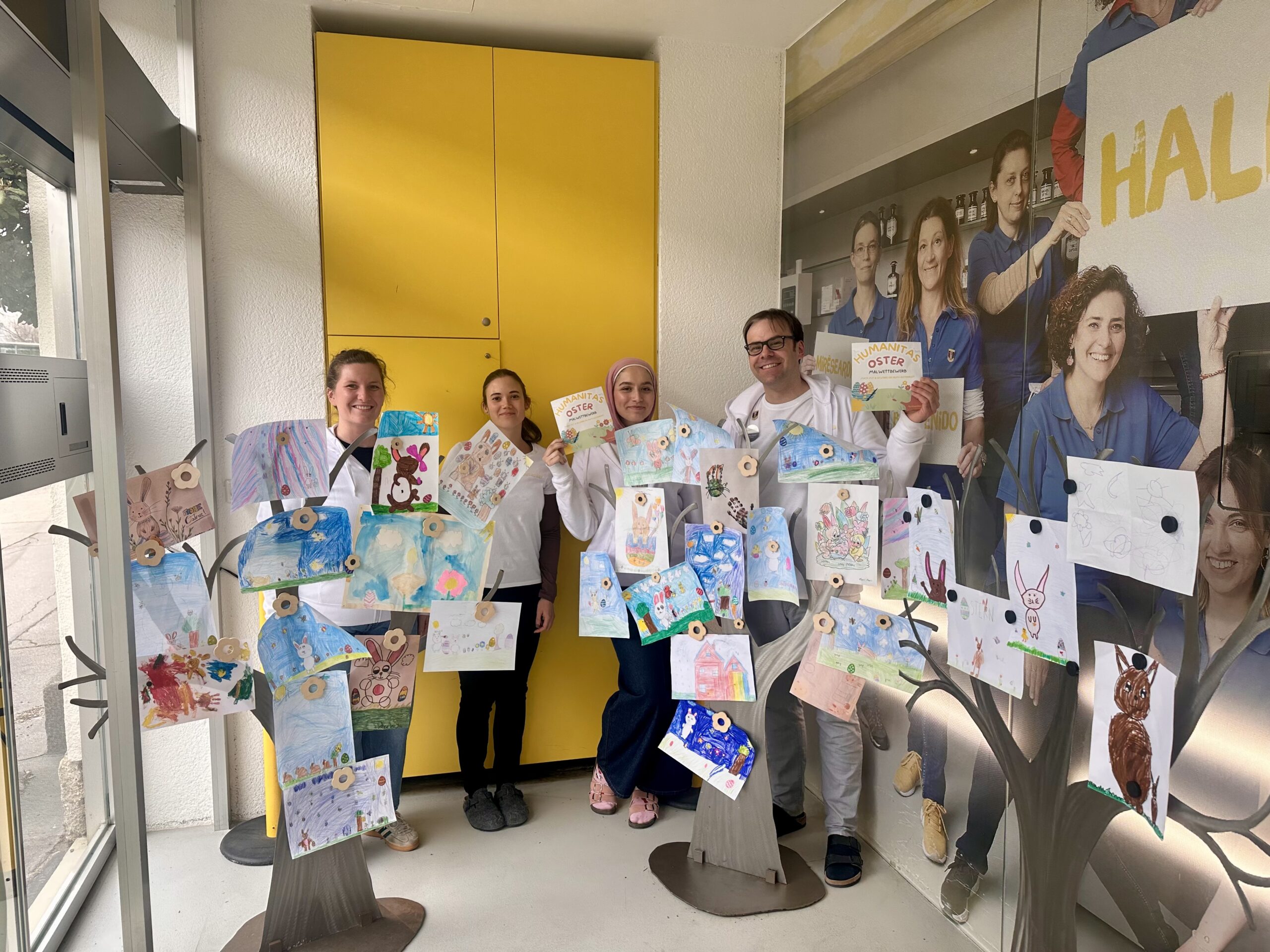 Das Apotheken Team präsentiert eine Oster-Kinderaktion mit bunten Zeichnungen von Kindern, die auf dekorativen Bäumen befestigt sind.