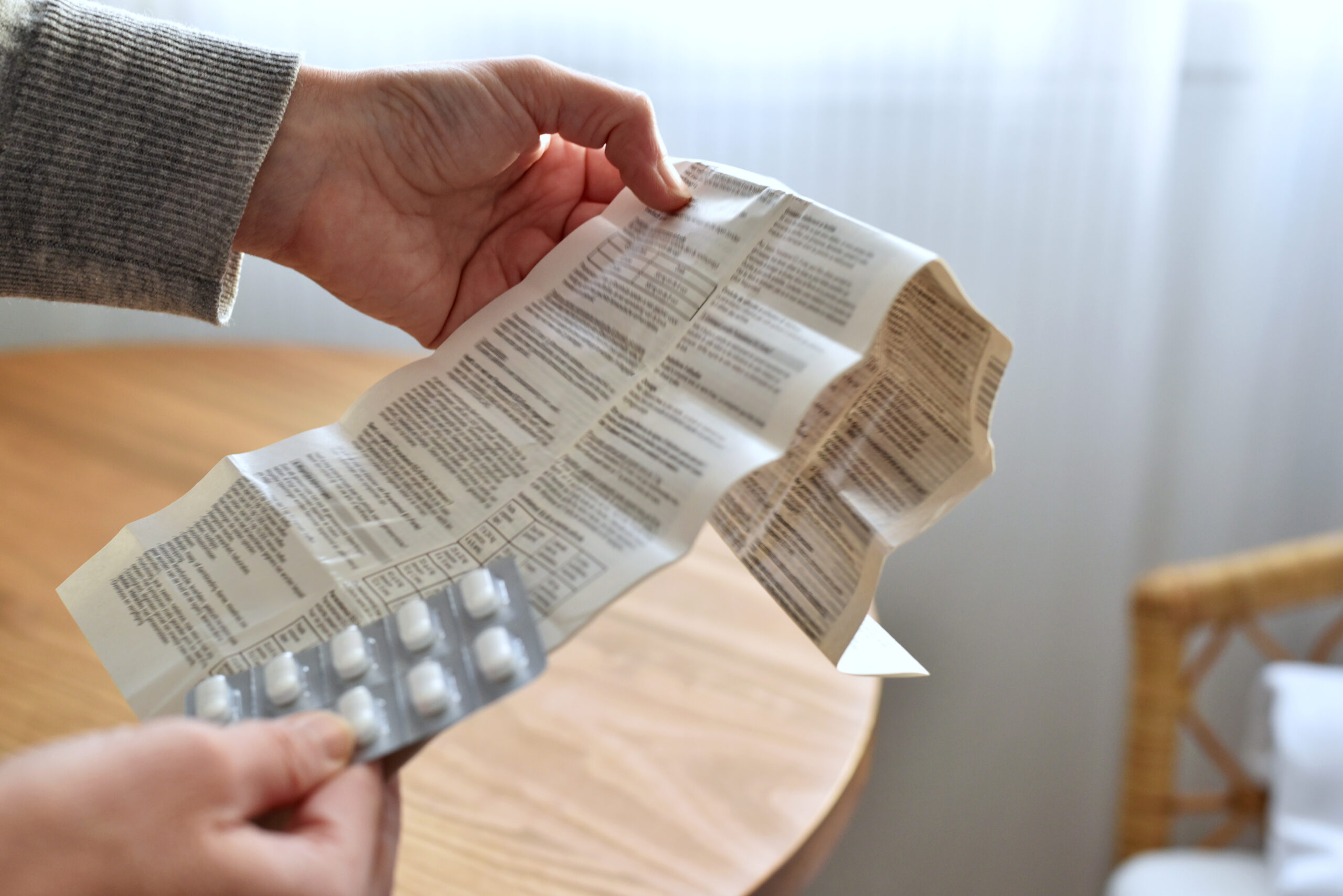 Symbolbild: Eine Hand haltet einen Beipackzettel und einen Tablettenblister in der Hand, im Hintergrund ist ein runder Tisch zu sehen.