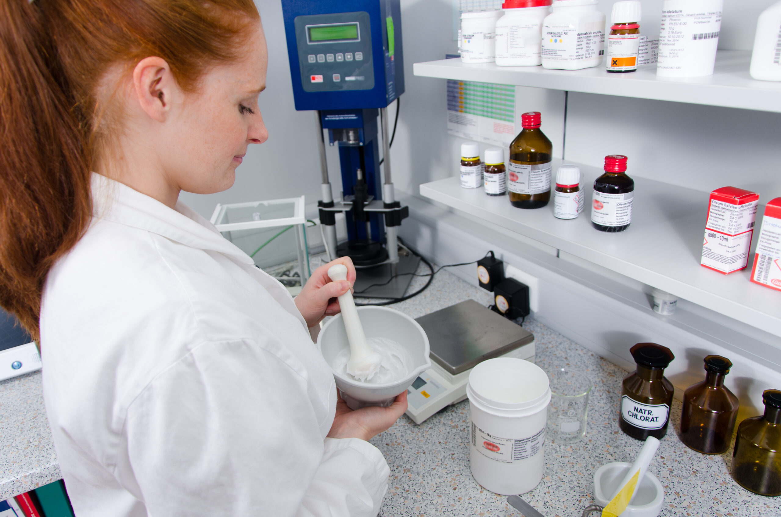 Symbolbild: Eine Apothekenmitarbeiterin steht im Rezepturbereich und verarbeitet mit einem Pistill eine weiße Masse in einer Patene. Im Hintergrund sind Waage, Geräte sowie Standgefäße auf Regalen zu sehen.