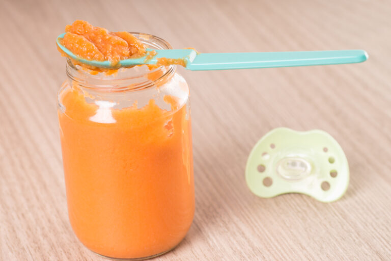 Symbolbild: Babygläschen mit orangem Inhalt und einem Löffel voll Nahrung darauf. Daneben liegt ein Schnuller.