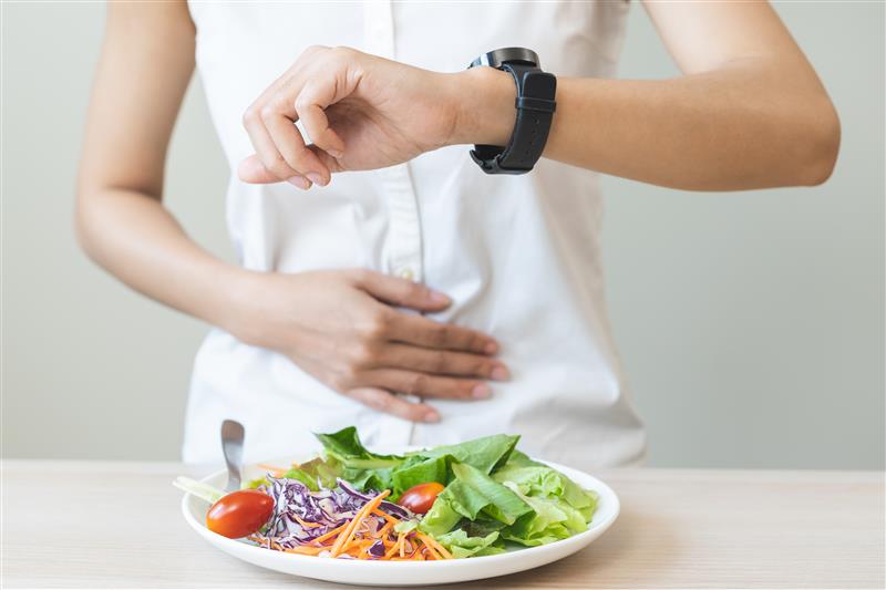 Symbolbild: Eine Frau steht vor einem Teller voll mit Gemüse der auf dem Tisch liegt, mit einer Hand greift sie sich auf den Bauch und schaut auf ihre Uhr am Handgelenk.