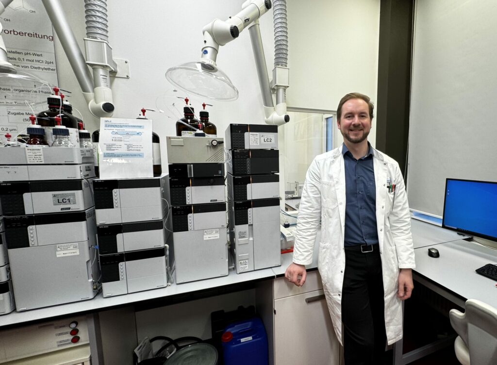 Der Laborleiter steht neben einer HPLC Anlage. Er trägt einen weißen Mantel und lächelt.