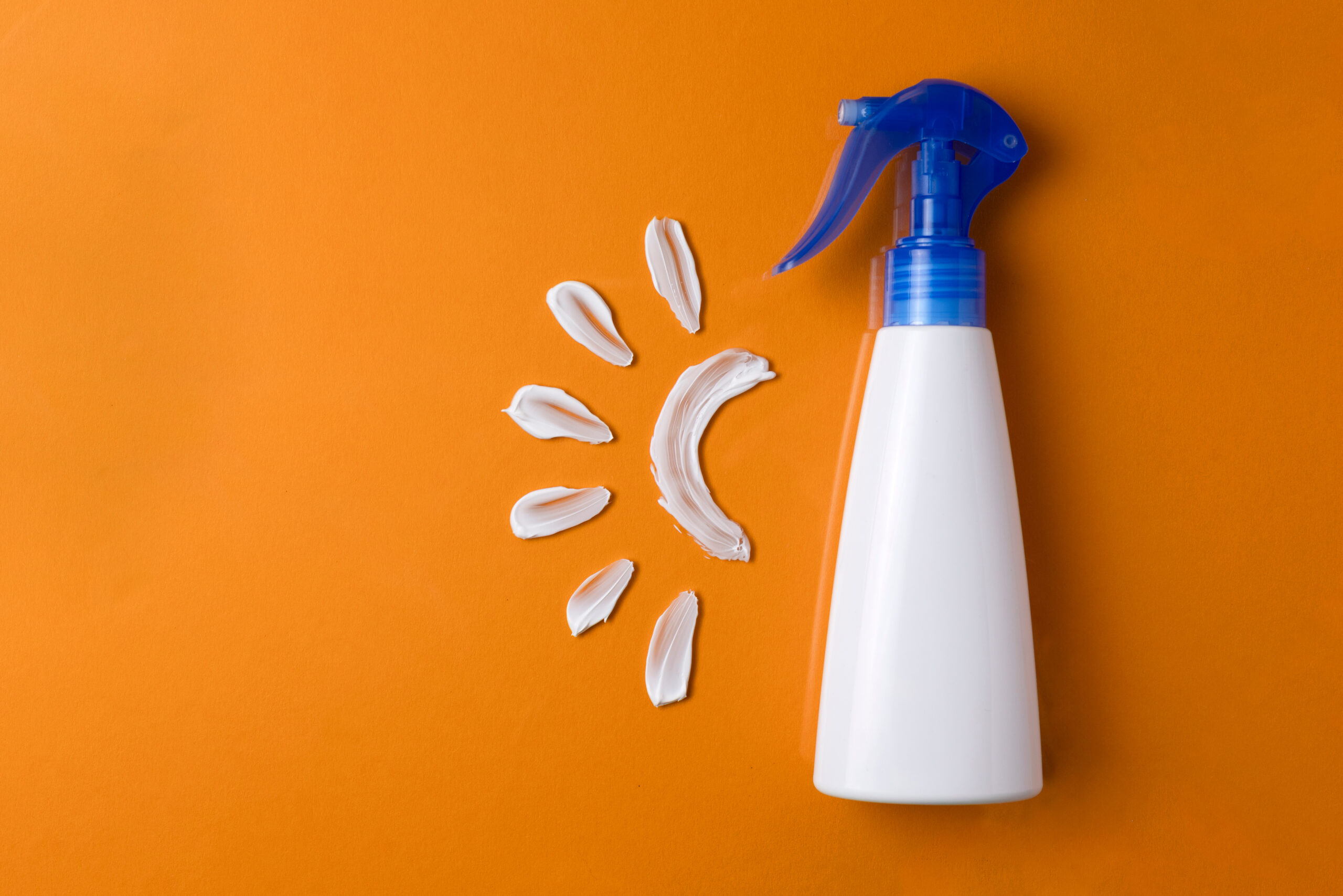Symbolbild: Weiße Sonnencremeflasche vor orangefarbenem Hintergrund. Mit Sonnencreme wurde daneben das Symbol einer Sonne gemalt.