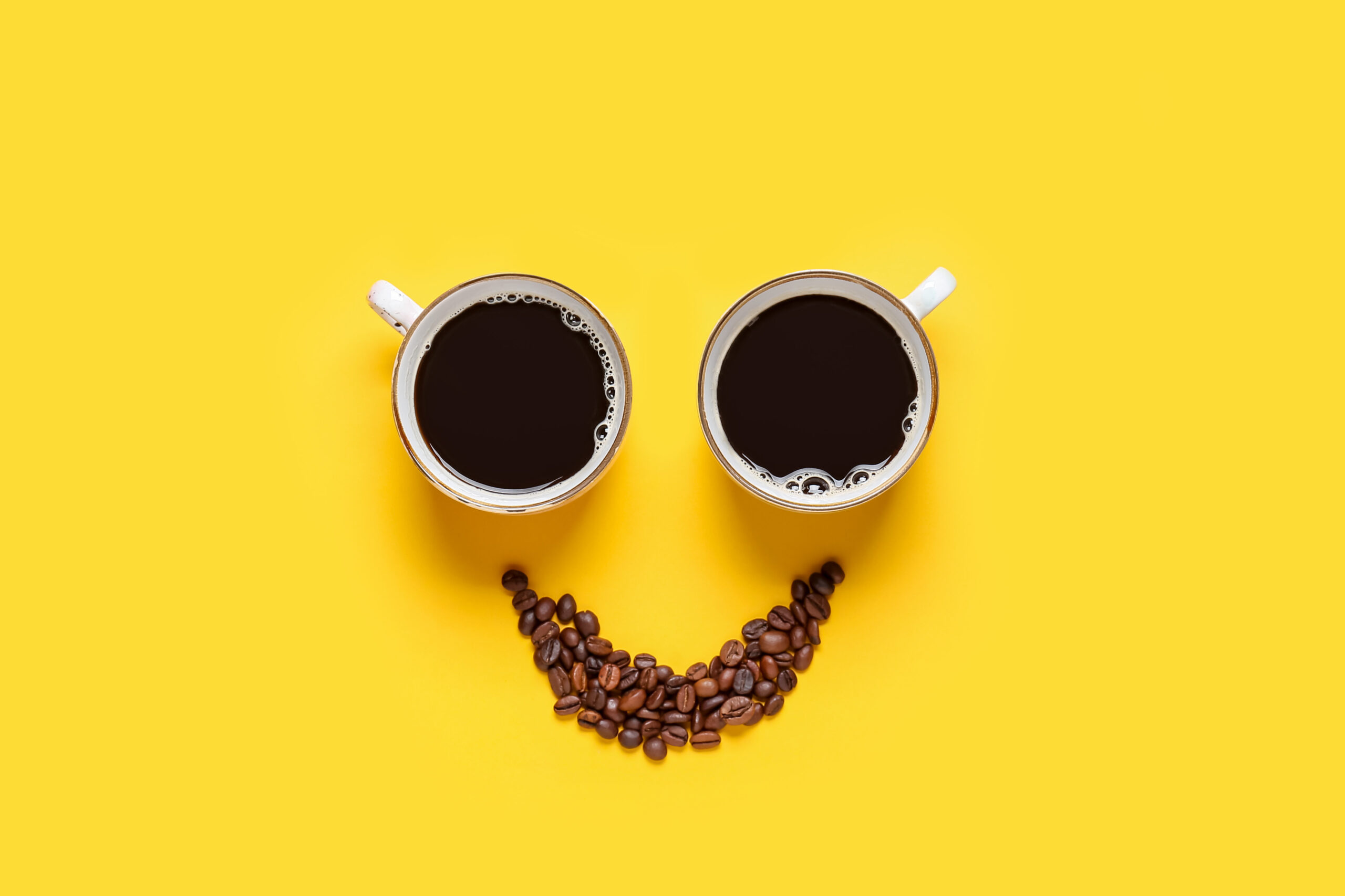 Symbolbild: Zwei Kaffeetassen und ein paar zusammen geschobene Kaffeebohnen auf einem gelben Untergrund.