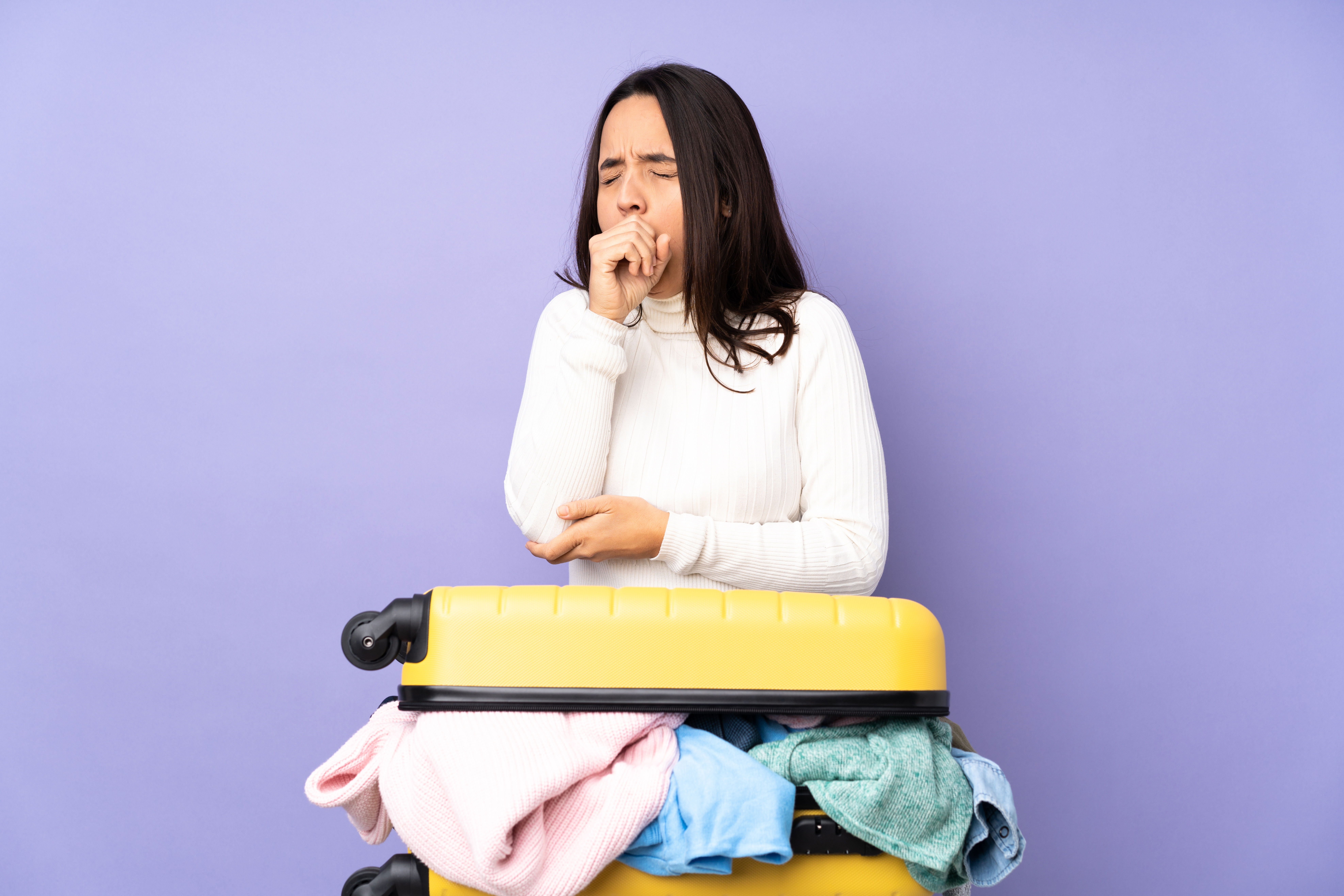Symbolbild: Eine Frau im weißen Pullover hustet und lehnt sich auf einen vollen Reisekoffer.