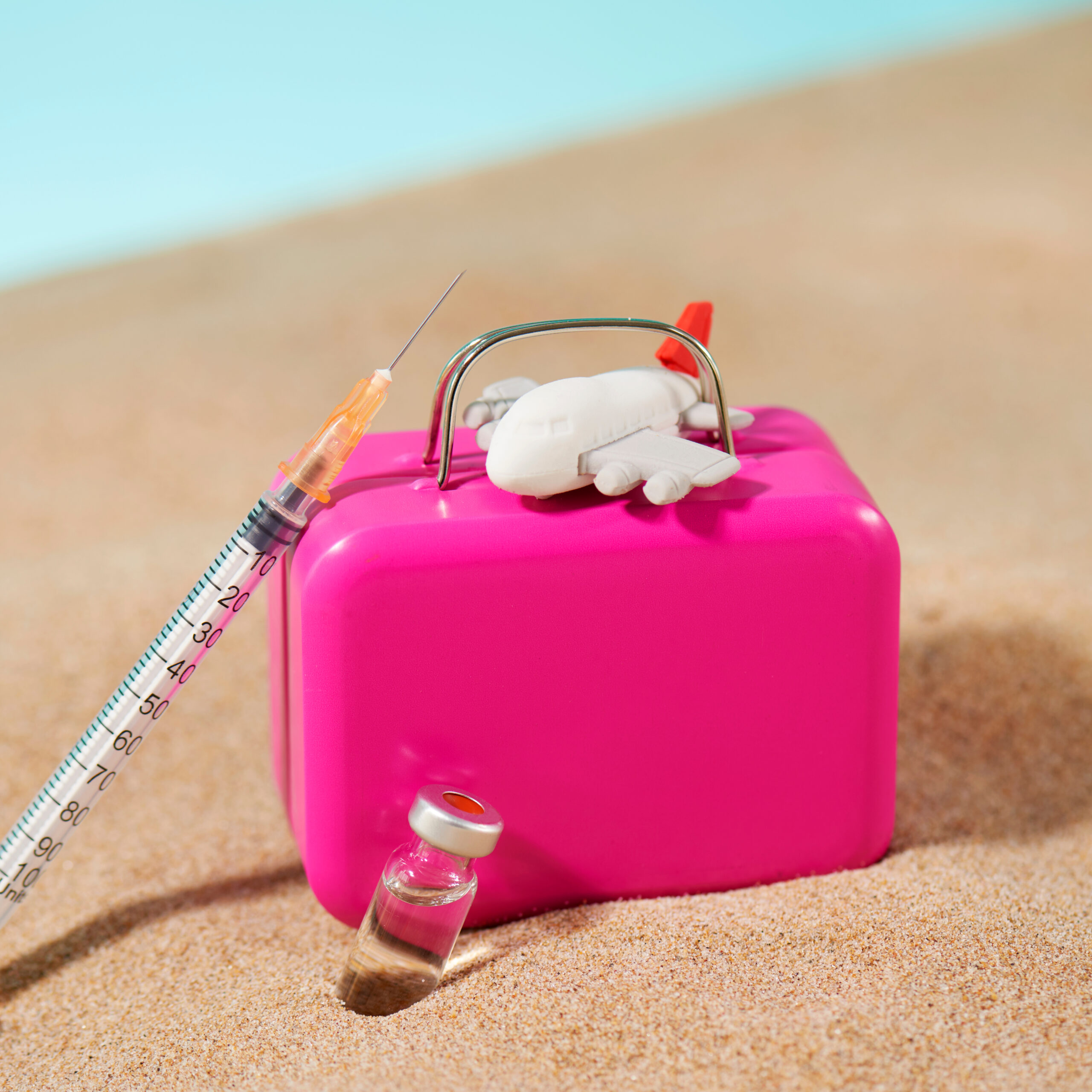 Symbolbild: Ein pinker Reisekoffer steht im Sand. Daneben lehnt eine Spritze und ein Flugzeug.
