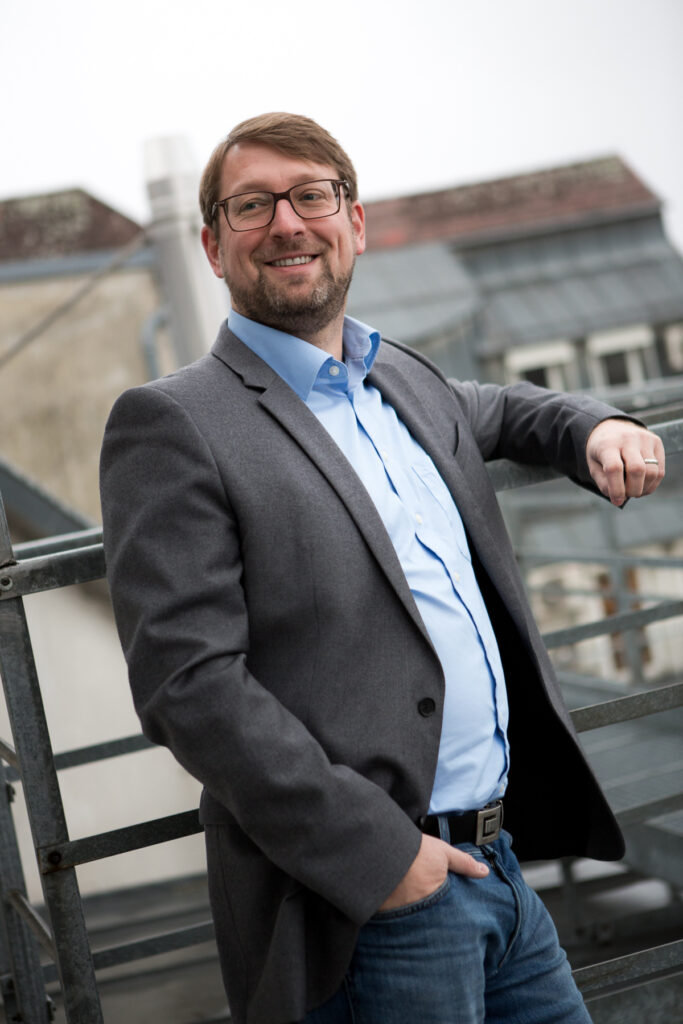 Mario Wolfran steht an einem Geländer. Er trägt einen Anzug und ein blaues Hemd.