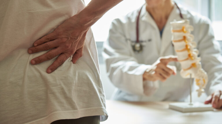 Symbolbild: Eine Patientin hält sich den unteren Rücken, während ein Arzt auf eine Wirbelsäulenmodell deutet.