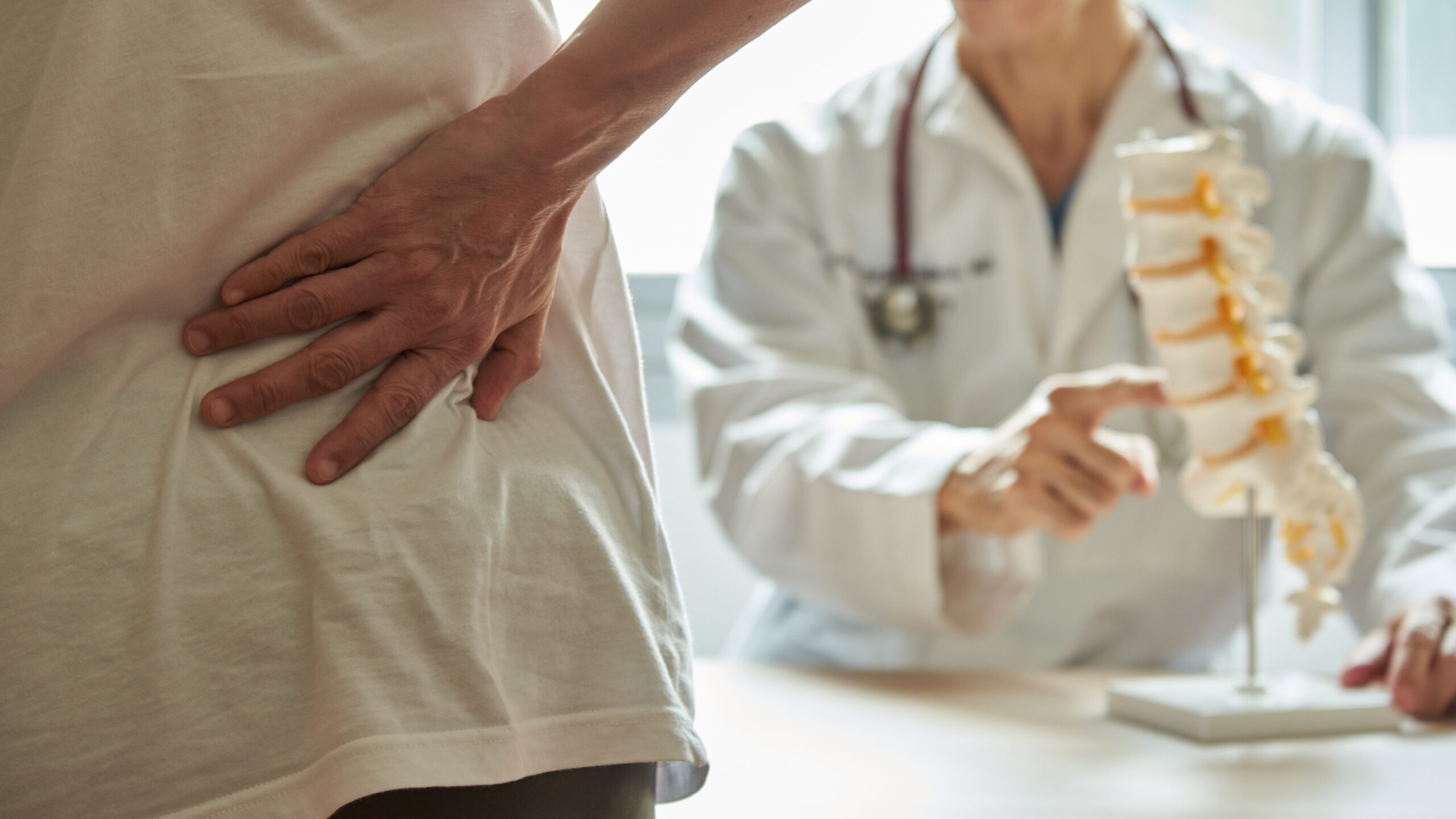 Symbolbild: Eine Patientin hält sich den unteren Rücken, während ein Arzt auf eine Wirbelsäulenmodell deutet.