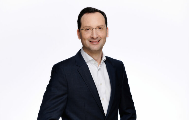 Julien Auger-Ottavi als Country President. Er trägt einen schwarzen Anzug und lächelt.